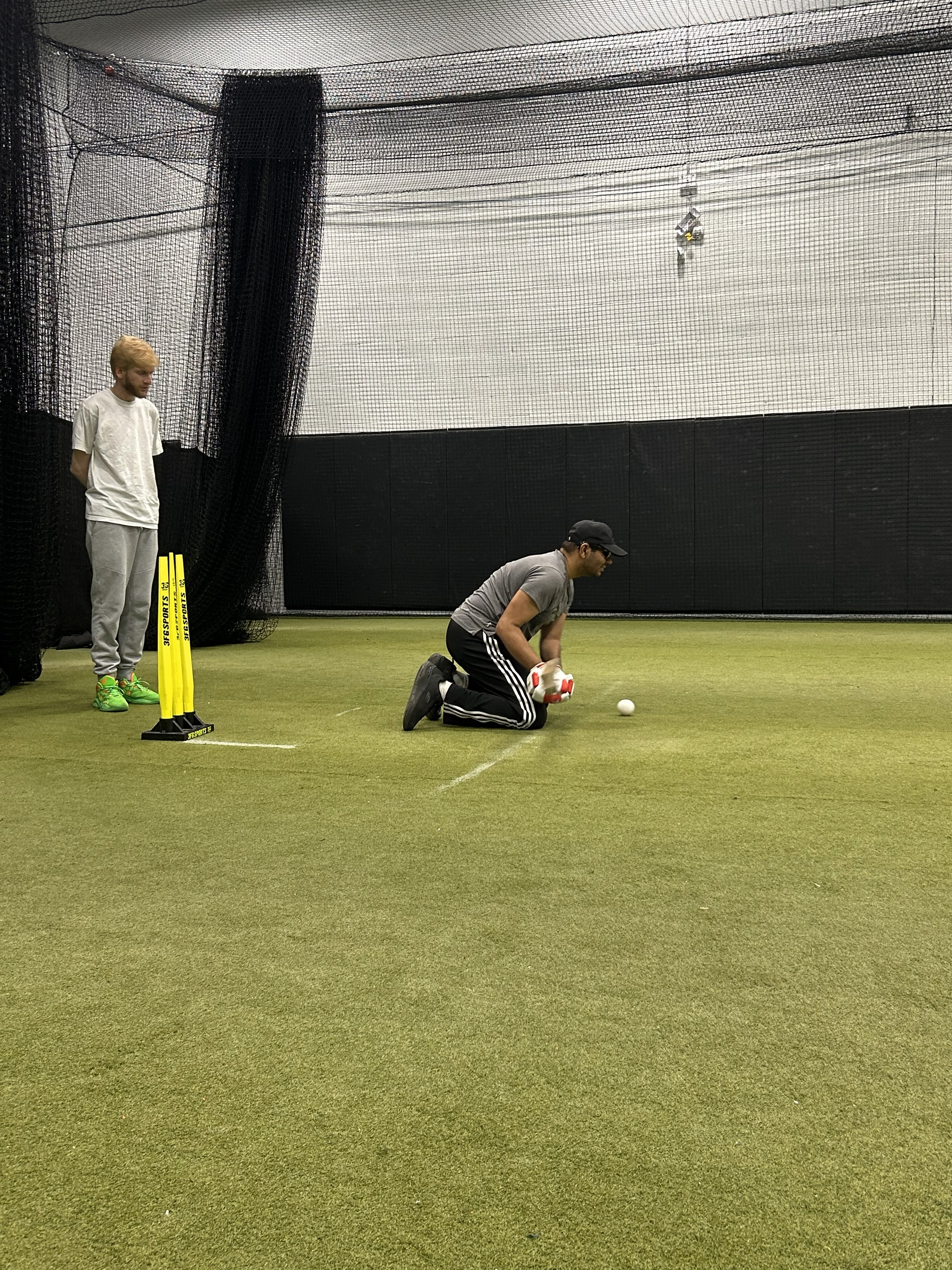 The batsman is kneeling on the indoor cricket pitch, preparing to play a shot at the ball in front of him, while the wicket-keeper stands slightly to the left behind the wickets, ready to react.